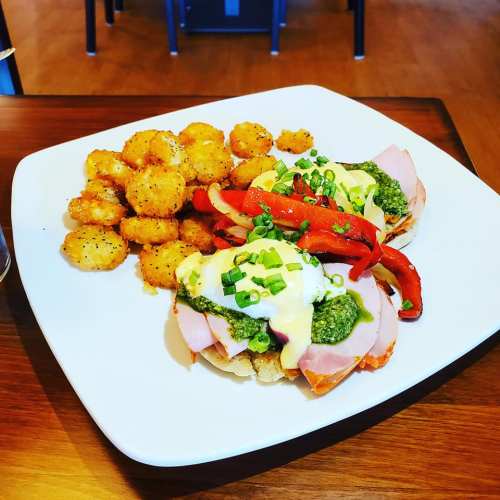 A plate of eggs Benedict with ham, pesto, and red peppers, served with crispy potato bites on the side.