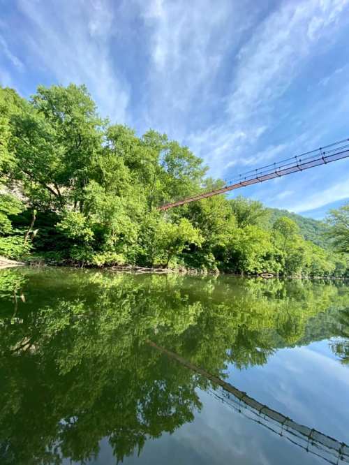 A serene river scene with lush green trees and a suspension bridge reflected in the calm water under a blue sky.