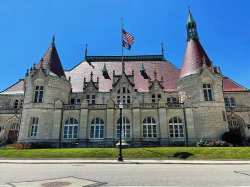 Historic building with pointed roofs, large windows, and an American flag in front, set against a clear blue sky.