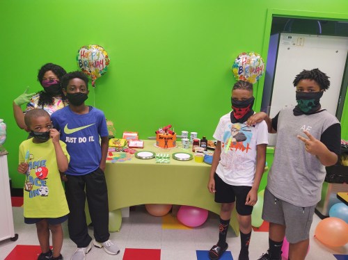 A group of children wearing masks stands by a birthday table decorated with balloons and treats in a colorful room.