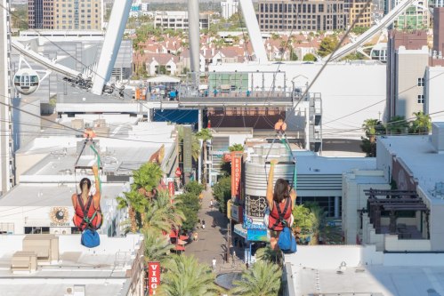 Two people ziplining above a vibrant street lined with shops and palm trees, with a large Ferris wheel in the background.
