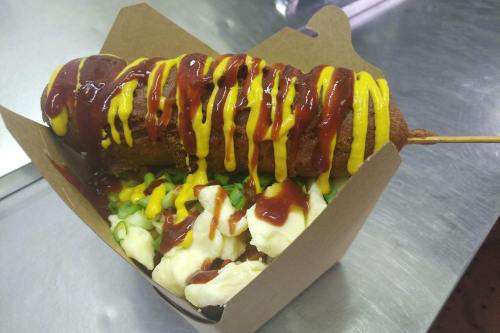 A corndog drizzled with mustard and ketchup, served with a side of mashed potatoes in a takeout box.