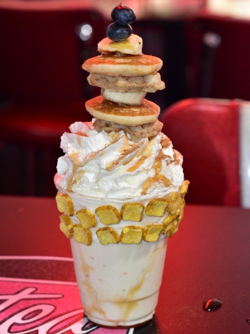 A tall milkshake topped with whipped cream, mini pancakes, blueberries, and cereal pieces, served in a glass.