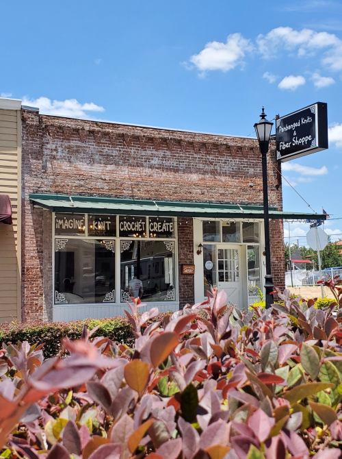 A cozy fiber shop with a brick exterior, featuring large windows and a sign that reads "Imagine Knit Crochet Create."