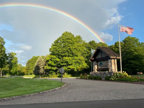 A scenic entrance sign with a rainbow overhead, surrounded by lush green trees and an American flag waving.