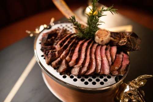 Sliced steak arranged on a decorative platter, garnished with herbs and a piece of roasted garlic.