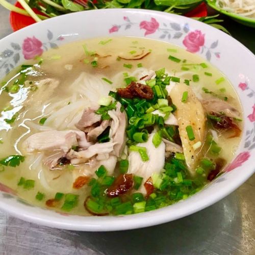 A bowl of chicken noodle soup with herbs, green onions, and sliced chicken in a clear broth.