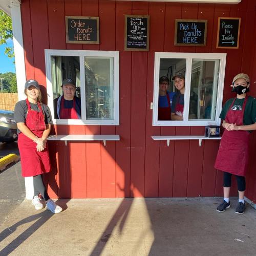 Four staff members in aprons stand at a donut shop window, with signs for ordering and pickup visible.