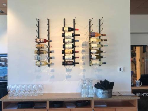 Three wine racks mounted on a wall display various bottles of wine, with glasses and carafes on a counter below.