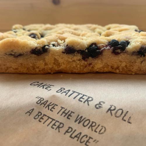 A close-up of a baked dessert with blueberries, resting on a parchment paper with the text "Bake the World a Better Place."
