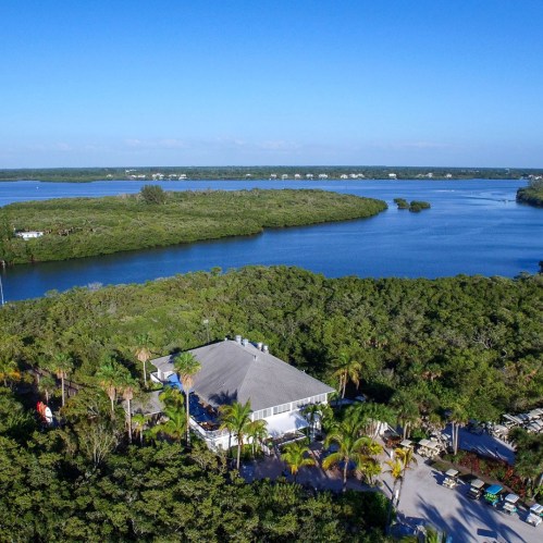 Aerial view of a waterfront property surrounded by lush greenery and a calm blue river under a clear sky.