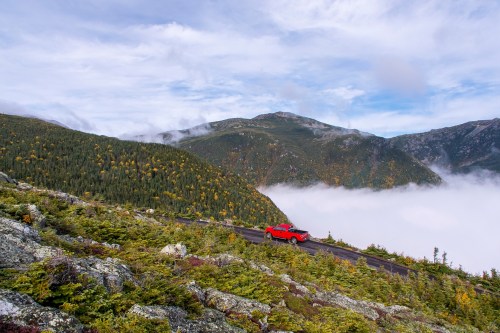 A red car drives along a winding road with mountains and fog-covered valleys in the background.