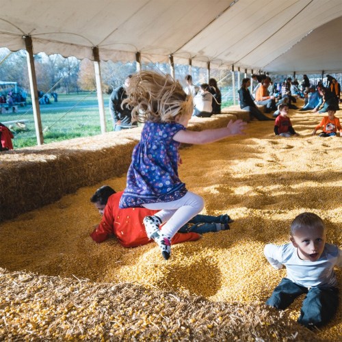 Children playing in a hay-filled area under a tent, with adults and other kids enjoying the outdoor event.