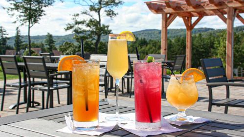 Four colorful cocktails on a table, with a scenic outdoor view of trees and mountains in the background.