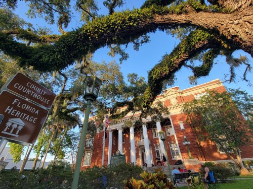 Historic county courthouse surrounded by lush trees and a sign for Florida Heritage. Clear blue sky above.