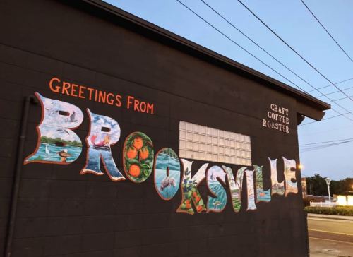 Colorful mural reading "Greetings from Brooksville" on a black wall, with "Craft Coffee Roaster" above.