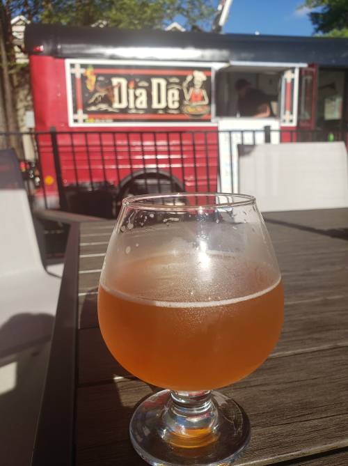 A glass of amber beer sits on a table, with a food truck in the background.