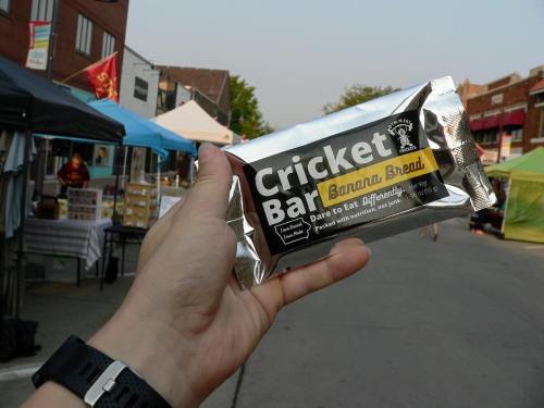 A hand holds a Cricket Bar labeled "Banana Bread" at an outdoor market with tents in the background.