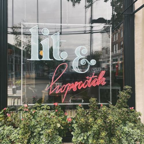 Neon sign reading "lit & hopscotch" in a storefront window, surrounded by greenery.