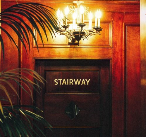 A wooden door labeled "STAIRWAY" with a chandelier above and a plant in the foreground.