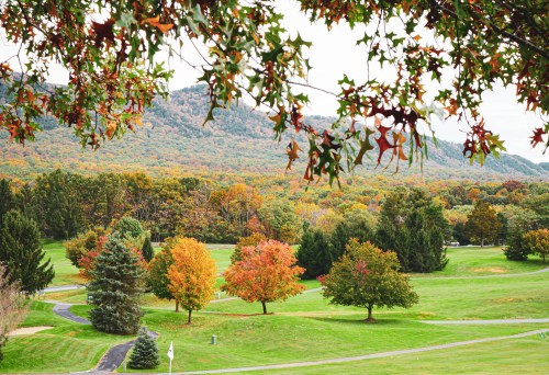 A scenic view of a golf course surrounded by trees with autumn foliage and mountains in the background.
