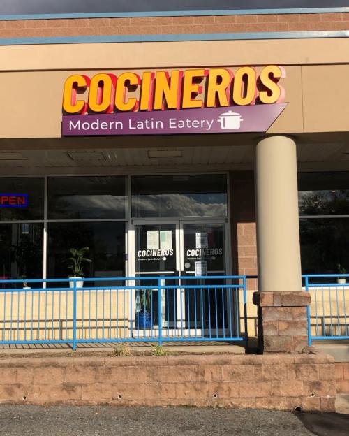 Sign for "Cocineros Modern Latin Eatery" on a storefront with a blue railing and open sign.