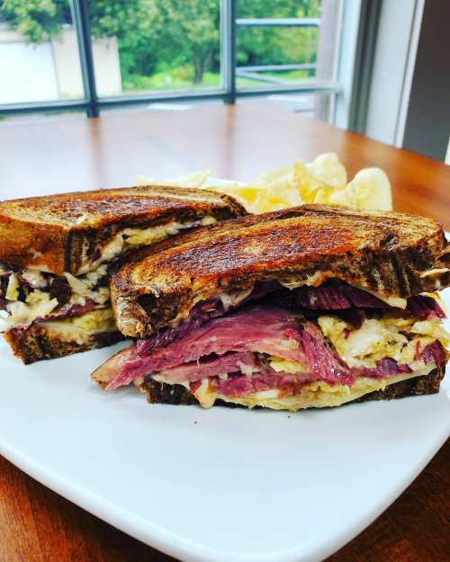 A close-up of a toasted sandwich filled with layers of meat and coleslaw, served on a white plate with chips.