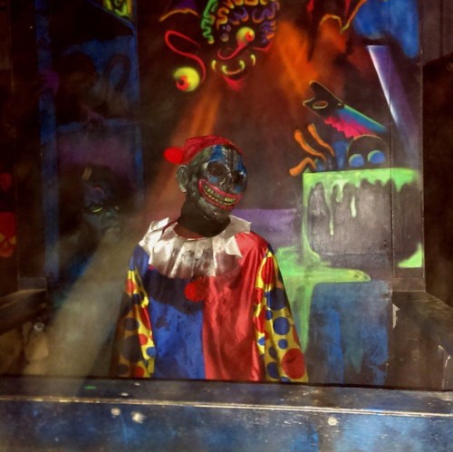 A creepy clown figure stands in a dark, colorful setting with glowing graffiti and eerie lighting.