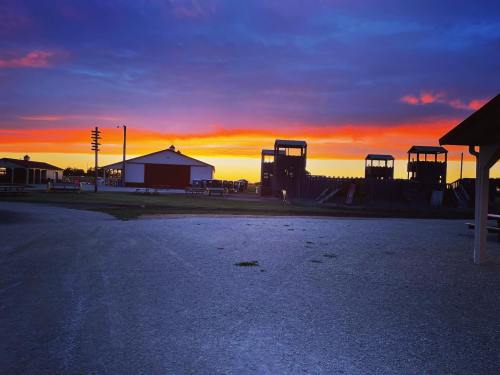 A vibrant sunset casts colorful hues over a rural landscape with buildings and structures silhouetted against the sky.