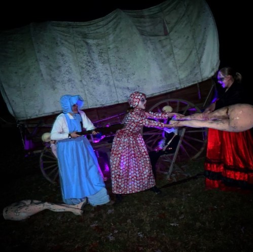 Three figures in costumes interact near a covered wagon, with eerie lighting and props creating a spooky atmosphere.