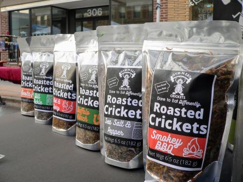 A row of colorful bags of roasted crickets, each labeled with different flavors, displayed on a table.