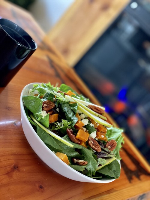 A fresh salad with greens, pecans, and squash, next to a black cup on a wooden table.