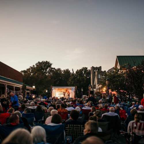 A large crowd gathers outdoors for a performance, with a stage and trees in the background during sunset.