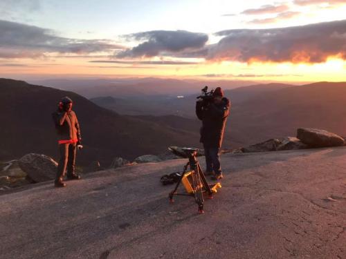 A person stands on a mountain at sunset while another films them with a camera on a tripod.