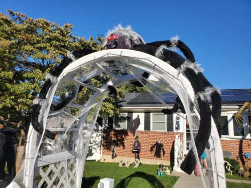 A white archway decorated with a large black spider and cobwebs, set in a yard with a clear blue sky.