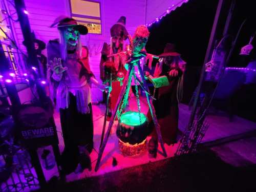 Three spooky figures in witch costumes gather around a glowing cauldron, surrounded by purple lights and Halloween decorations.