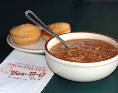 A bowl of chili with a spoon, accompanied by two cornbread muffins and a napkin from Moonlite Bar-B-Q Inn.