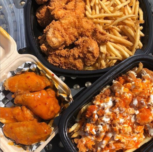 A variety of fried foods: crispy chicken, French fries, buffalo wings, and loaded fries in takeout containers.