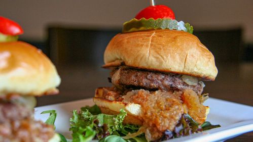 A close-up of a juicy burger with lettuce, pickles, and a cherry tomato on a toasted bun, served on a plate.