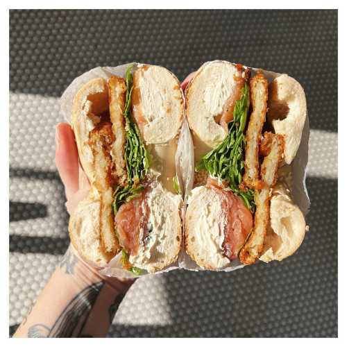 A hand holding a large sandwich with layers of lettuce, tomato, and crispy fried items, cut in half.