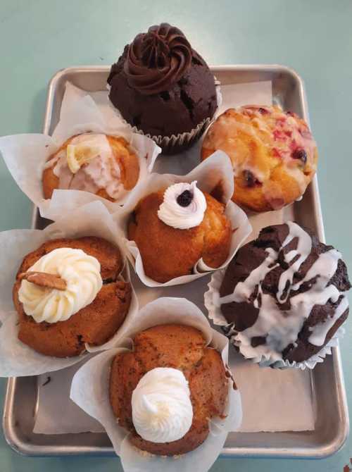 A tray of assorted muffins, including chocolate, lemon, and fruit varieties, each topped with frosting or cream.