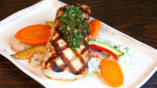 Grilled fish fillet topped with green sauce, served with colorful vegetables and rice on a white plate.