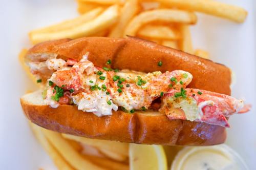 A lobster roll topped with chives, served with French fries and a side of lemon and dipping sauce.