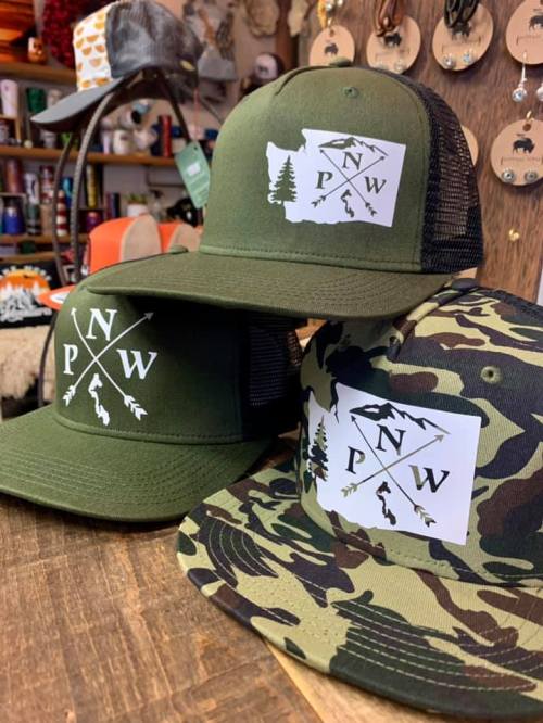Three hats featuring a PNW logo, one in olive green, one in camouflage, displayed on a wooden surface.