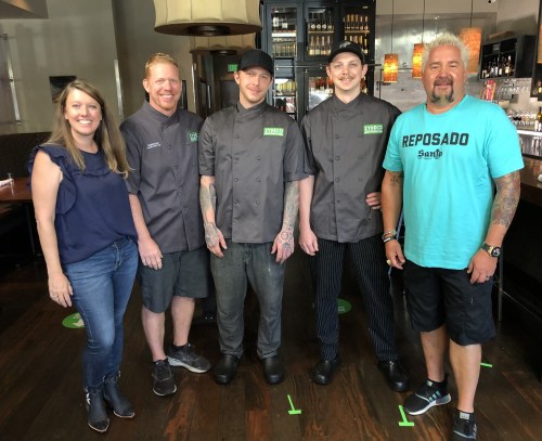 A group of five people, including chefs in uniforms and a woman, stand together in a restaurant setting.