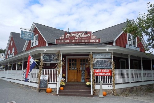 A charming red building with a porch, featuring signs for a café and ATM, decorated with pumpkins for fall.