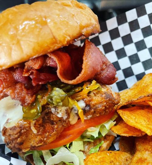 A close-up of a burger with crispy bacon, pickles, lettuce, and tomato, served with a side of potato chips.