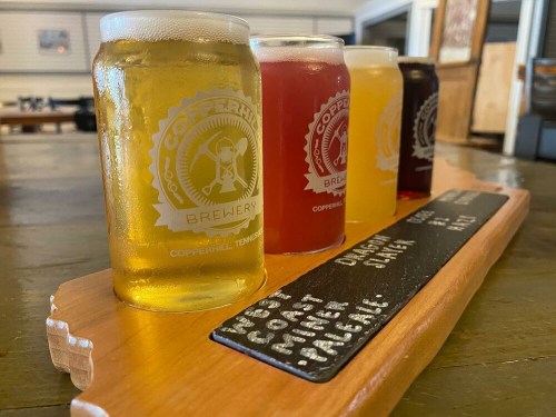 Four craft beer samples in glasses on a wooden flight board, with a brewery logo and tasting notes visible.