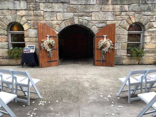 A stone archway with wooden doors, flanked by floral arrangements, leading to a wedding ceremony setup with white chairs.