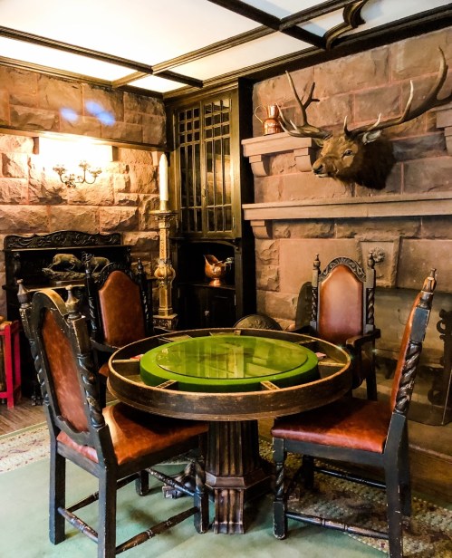 A cozy room featuring a round table with a green top, surrounded by wooden chairs and a stone fireplace with a mounted deer head.